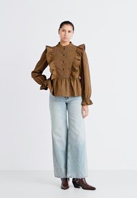 Blusa marrone a fantasia con dettagli in volants, chiusura a bottoni, e vita peplum, abbinata a jeans a gamba larga azzurri e stivaletti marroni.