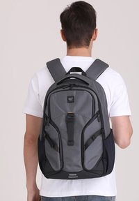 Grijze rugzak met zwarte accenten, gestructureerde stof, gewatteerde schouderbanden, meerdere ritsen en zijvakken. Ontworpen voor comfort en functionaliteit.
