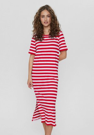 Ung kvinde med krøllet hår iført en kortærmet midi-kjole i rød og hvid vandret stribet med en sideslids, stående foran en ensfarvet baggrund.