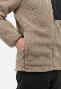 Personne portant une veste en polaire beige avec une main dans une poche latérale, associée à un pantalon noir, sur un fond blanc.