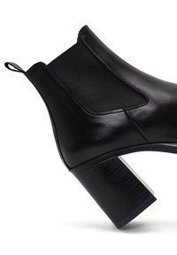 Bota de tobillo de cuero negro con punta redondeada, paneles laterales elásticos y un tacón de bloque de madera texturizada. Presenta un acento plateado en contraste.