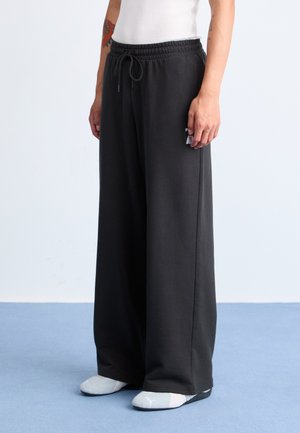 Black wide-leg sweatpants with elastic waistband, drawstring, and side pocket. Soft texture, worn with gray slip-on shoes.