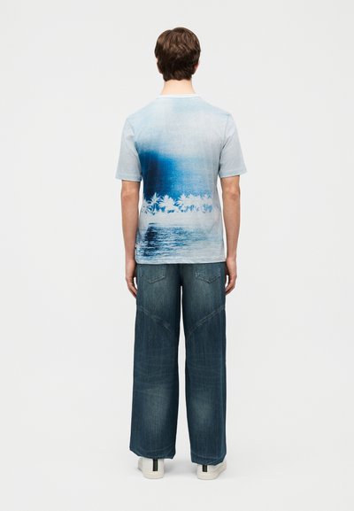 Άνδρας που στέκεται με την πλάτη στην κάμερα, φοράει μπλε μπλουζάκι ομπρέ με σχέδιο φοίνικα και τζιν παντελόνι φαρδύ με λευκά αθλητικά παπούτσια.