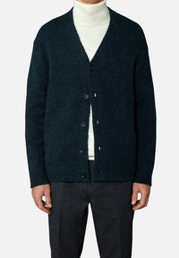 Donkergroene cardigan gemaakt van gestructureerde gebreide stof, met een V-hals, knoopsluiting en geribde manchetten en zoom. Gedragen over een witte coltrui.