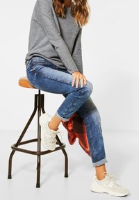 Jean en denim bleu avec les ourlets retroussés, associés à une chemise à manches longues grise et des baskets blanches, assis sur un tabouret en métal. Détail de siège en cuir marron.