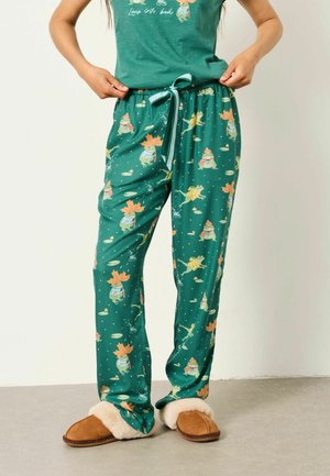 Person trägt grüne Pyjamahosen mit Frosch- und Herbstlaub-Druck, kombiniert mit braunen Hausschuhen mit weißem Fellfutter.