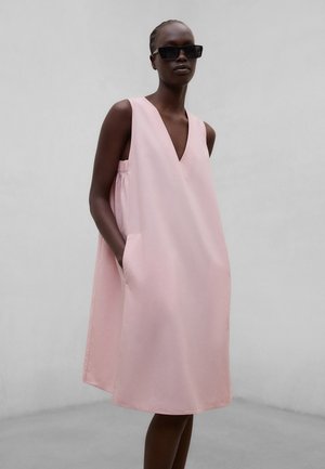 Kvinne iført ermeløs, løs og lys rosa kjole med dyp V-hals og sidelommer, accessorized med store svarte solbriller.