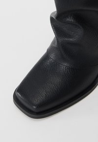 Botte en cuir noir avec un bout carré et une surface texturée, dotée d'une partie supérieure douce et froncée ainsi que d'une semelle plate.