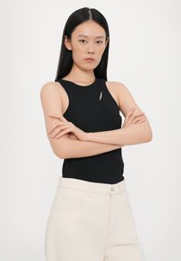 Black sleeveless top with a rounded neckline and a cutout detail. Worn with high-waisted light beige pants made of denim fabric.