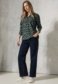Camicia verde a maniche lunghe con motivo zigzag, abbinata a jeans a gamba larga blu scuro e sneakers bianche. Tessuto morbido con design con colletto.