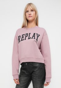 Šviesiai rožinė trumpa džemperis su juodu spausdintu „REPLAY“ logotipu, su gipiūrinėmis rankovėmis ir apačia, derinama su juodomis tekstūruotomis kelnėmis.