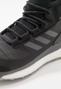 Zapatilla deportiva negra de Adidas con parte superior texturizada, tres franjas reflectantes, agujetas negras y suela acolchada blanca sobre un fondo blanco.