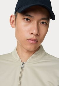 Beige bomberjack met een geribbelde kraag, voorzien van een ritsluiting en een gladde textuur. Donkere pet recht naar voren gedragen, minimale gezichtsuitdrukking.