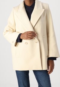 Cappotto doppio petto in misto lana crema con rever a rovescio, tasche laterali e dettagli con bottoni, abbinato a un maglione blu navy e jeans.