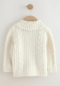 Suéter blanco de punto con un cuello alto y patrones de cable texturizados. Presenta puños y dobladillo acanalados, colgado en un perchero de madera.