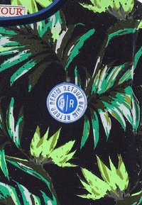 Tissu noir avec des motifs de feuilles vertes et vert clair. Présente unePatch logo ronde bleue étiquetée "RETOUR DENIM". Matériau texturé et extensible.