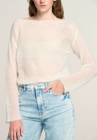 Maglione leggero e trasparente in un morbido color crema, con una vestibilità ampia, scollo largo e maniche lunghe. Abbinato a jeans azzurri a vita alta.