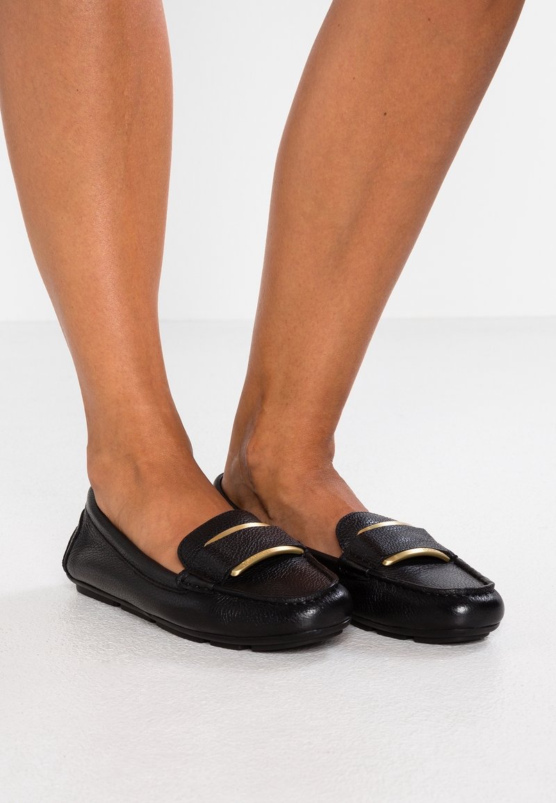 Mocassins en cuir noir avec finition texturée et accents dorés. Bout arrondi et semelle flexible. Présentés sur deux pieds, debout sur une surface blanche.