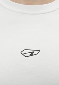 Κοντινή λήψη ενός άσπρου μπλουζιού με λαιμόκοψη, που διαθέτει ένα μικρό μαύρο γεωμετρικό λογότυπο στο κέντρο του στήθους.