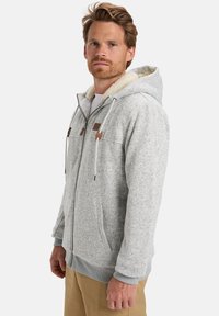 Muž v svetlo šedej zipsovej mikine s fleece podšívkou a šnúrkami, v kombinácii s béžovými nohavicami, stojí na neutrálnom pozadí.