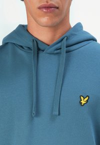 Primo piano di una persona che indossa una felpa blu con cappuccio e cordini, con un piccolo logo di un uccellino giallo sul petto.
