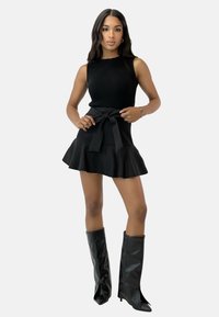 Top negro de canalé sin mangas combinado con una falda corta negra acampanada que presenta un lazo en la cintura; la modelo lleva botas altas brillantes negras.