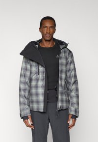 Chaqueta de hombre a cuadros en tonos gris y verde, con cierre de cremallera, capucha ajustable y múltiples bolsillos. La tela tiene una textura suave y resistente al agua.