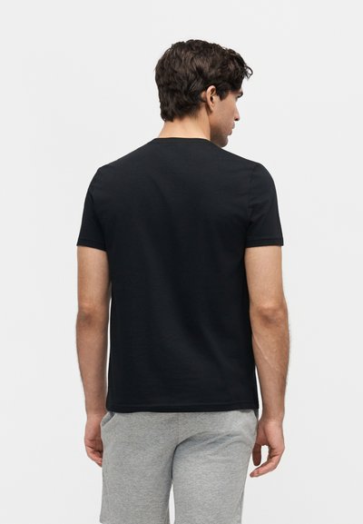 Άνδρας με σκούρα μαλλιά που φορά απλό μαύρο κοντομάνικο μπλουζάκι και ανοιχτό γκρι σορτς, με την πλάτη γυρισμένη σε λευκό φόντο.