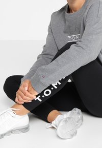 Persoon in een grijze Tommy Hilfiger sweatshirt en zwarte leggings met witte sneakers, zittend met gekruiste benen op een witte vloer.