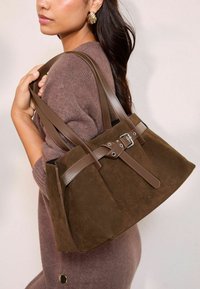 Pruunest nahast kott, millel on nahkrihmad, funktsiooniks on pandla detail ja õmblusaktsendid. Koti kuju on struktureeritud.