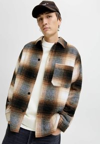 Button-up shirt with a plaid pattern in browns, whites, and blues; loose fit, large chest pocket, worn over a white sweater.