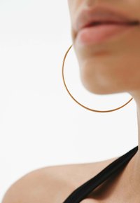 Pendiente de aro dorado, grande y redondo, con un acabado metálico suave. Diseño minimalista, llevado en la oreja de una persona, contra un fondo neutro.