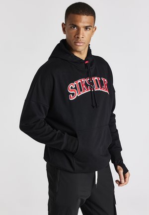 La sudadera negra cuenta con un logo bordado en rojo, cordones ajustables y un bolsillo frontal. El material es suave y texturizado, con puños acanalados.