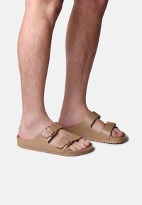 Tan slip-on sandaler med två justerbara spännen, en strukturerad sula och en lätt gummisula.