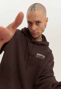 Brown hooded sweatshirt with a front pocket and "COFFEE HARMONY" logo. Material appears soft with a relaxed fit. Accented by drawstrings.