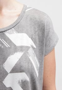 Šedé tričko s krátkým rukávem a volným střihem, zdobené bílým geometrickým vzorem a měkkou, texturovanou tkaninou. Kolo výstřihu.