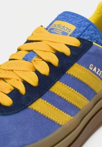 Τα αθλητικά παπούτσια Adidas Gazelle διαθέτουν μπλε σουέντ υλικό, κίτρινες υφασμάτινες κορδόνια, τρεις κίτρινες ρίγες και ταμπά λαστιχένια σόλα. Περιλαμβάνονται μοναδικές διακοσμητικές λεπτομέρειες με σχέδιο.