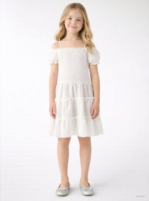 Giovane ragazza in piedi, indossa un vestito bianco smock con spalle scoperte, maniche a sbuffo e ballerine argento, su sfondo neutro.