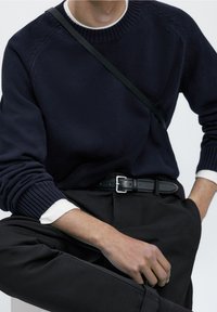 Personne portant un pull marine sur une chemise blanche, un pantalon noir, une ceinture noire et une sangle noire en bandoulière, assise avec une main dans la poche.