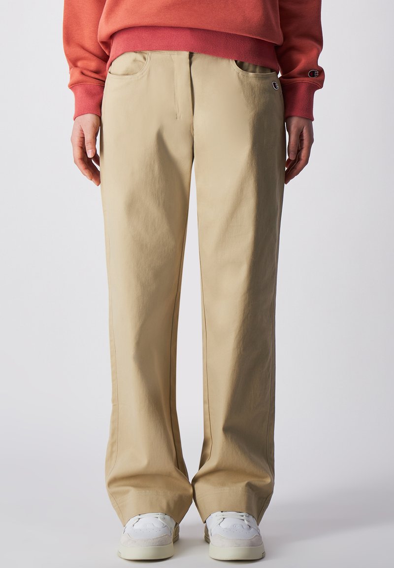 Beige weite Baumwollhosen mit Seitentaschen, einer Vordernaht und subtilen Nähdetails, kombiniert mit weißen Sneakers.