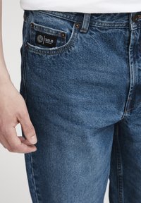 Denimjeans i mørk blå vask med svake slitasjemerker, med en fremre lomme og et teksturert merke sydd på linningen.
