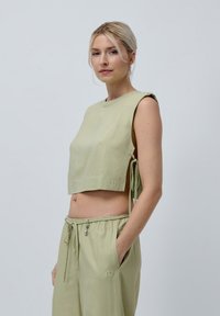 Korte mouwloze top van lichtgroene stof met zijsstrikjes en een zakdetail aan de voorkant, gecombineerd met bijpassende lichtgroene wijde broek.