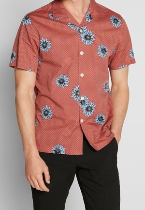 Homme portant une chemise à manches courtes couleur rouille avec de grandes impressions florales bleues et de petits motifs géométriques, associée à un pantalon noir.