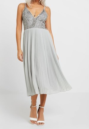 Kobieta w srebrnej, bezrękawnikowej bluzce z cekinami, plisowanej jasnoszarej spódnicy midi oraz białych szpilkach z paskiem wokół kostki.