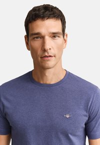 Mand med kort mørkt hår iført en ensfarvet blå rundhalset T-shirt med et lille GANT-logo på venstre bryst mod en hvid baggrund.