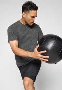 Herre treningsantrekk med en teksturert grå kortermet skjorte og svarte shorts, som holder en matt svart medisinball.