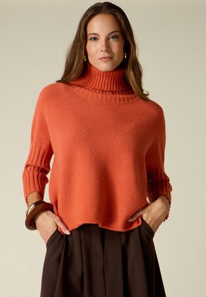 Mujer con un jersey de cuello alto naranja con las mangas remangadas y pantalones marrón oscuro, con las manos en los bolsillos, fondo neutro.