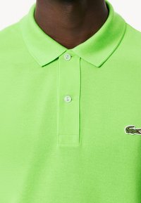 Osoba s tmavou pletí, oblékající jasně zelené polo tričko s límečkem, se zapnutými dvěma knoflíky a malým krokodýlím logem na hrudi.