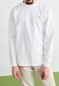 Man in een witte lange mouwen shirt met een klein geel logo op de borst en beige broek, staande op een groen en wit vloerpaneel.