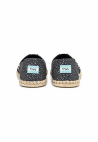 Sapatos slip-on de tecido preto com bolinhas brancas. Apresenta uma sola de espadrilha em juta e um rótulo azul claro com "TOMS" no calcanhar.